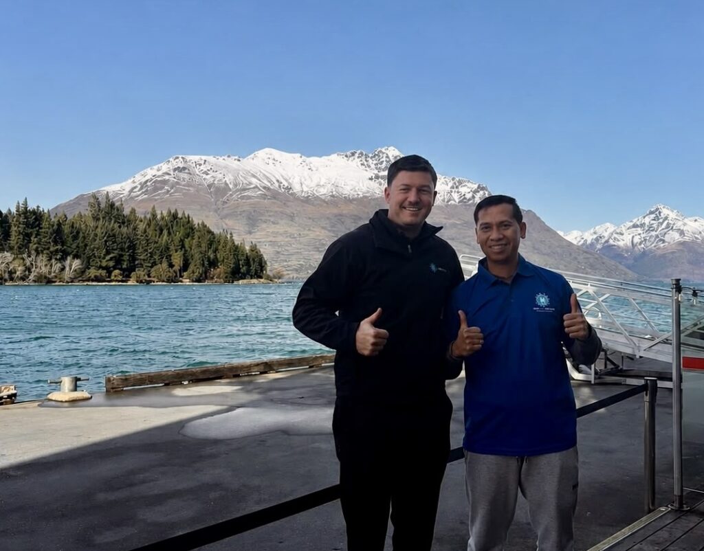 Two New Era team members standing beside Lake Wakatipu in Queenstown, New Zealand, with snow-covered mountains behind them