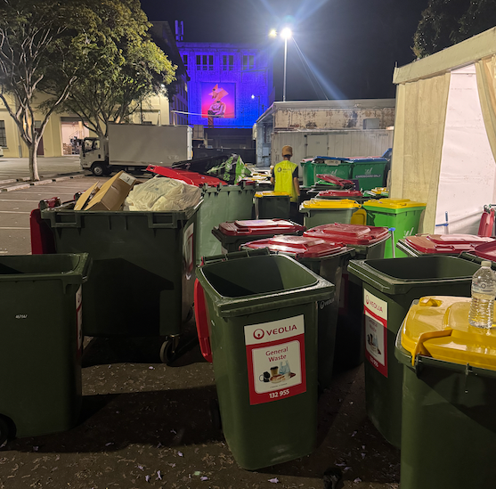 New Era team member in a high-visibility vest working among colour-coded event bins on a street beside the illuminated Night Feast venue at night