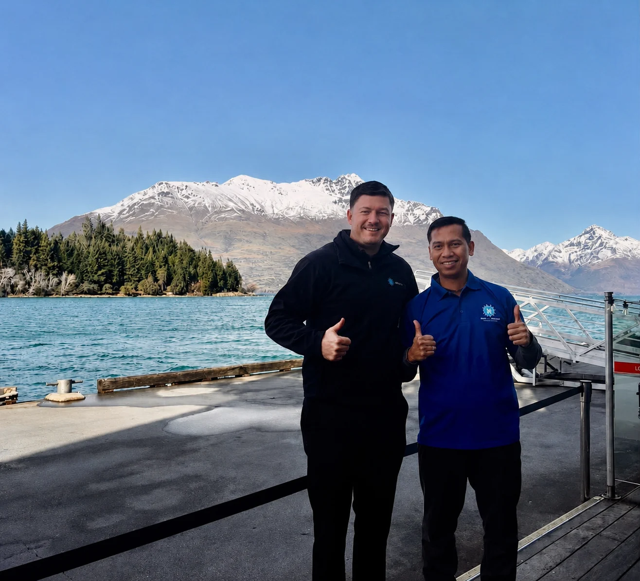 Two New Era team members in branded uniforms standing on a Queenstown lakefront wharf with Lake Wakatipu and snow-capped mountains behind them
