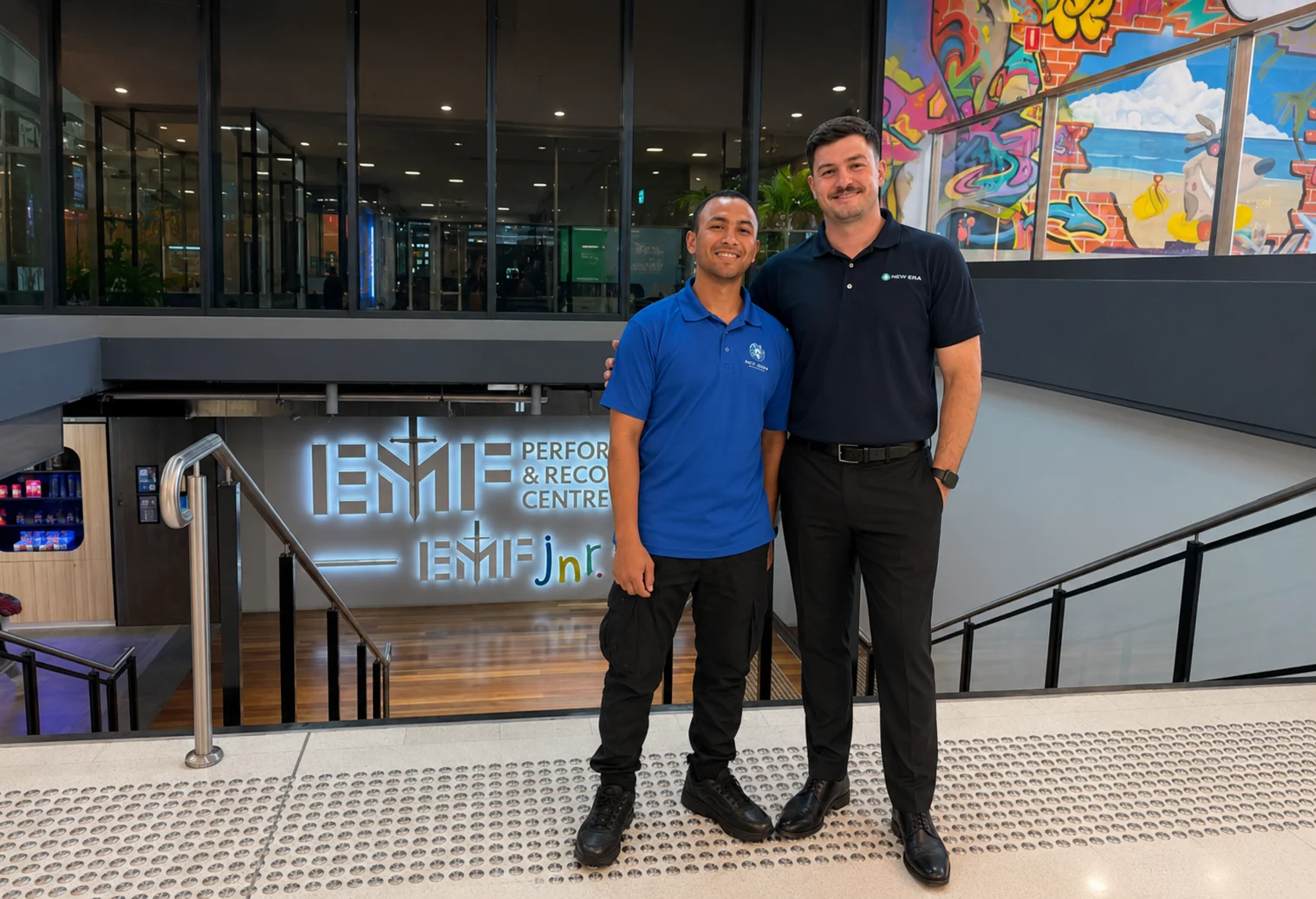 A New Era account manager and site team member in branded polos inside the entry atrium of a modern commercial performance centre