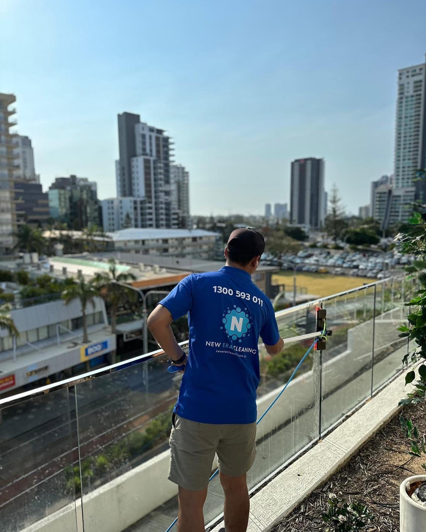 New Era team member in a branded polo cleaning balcony glass balustrades with a water-fed pole above a city outlook