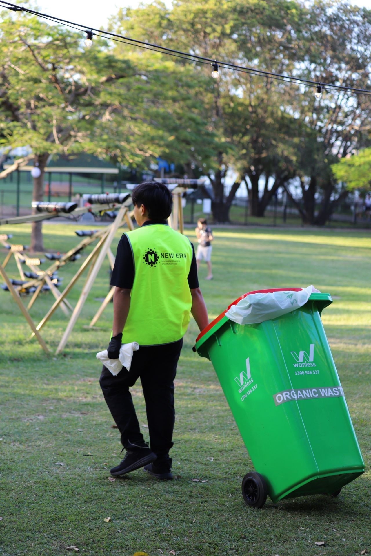 New Era team member in a New Era high-visibility vest wheeling a green organic waste bin across the Night Feast event lawn